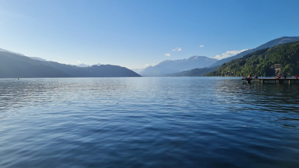 Der Millstätter See von Osten aus mit einer ruhigen, leicht bewegten Oberfläche, im Hintergrund die sanften Berge, eine beruhigende Atmosphäre geht von der Szenerie in blauen Tönen aus.